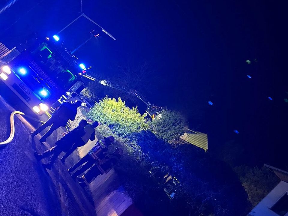 Foto zum Einsatzbericht: 022026 Dachstuhlbrand in Scheuerfeld, abgebildet sind Einsatzkräfte der Feuerwehr und DRK, sowie ein Fahrzeug der Feuerwehr